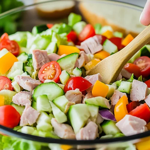 A fresh and colorful Sub in a Tub recipe in a glass bowl, featuring chopped lettuce, diced turkey, cucumbers, cherry tomatoes, and bell peppers, being mixed with a wooden spoon.